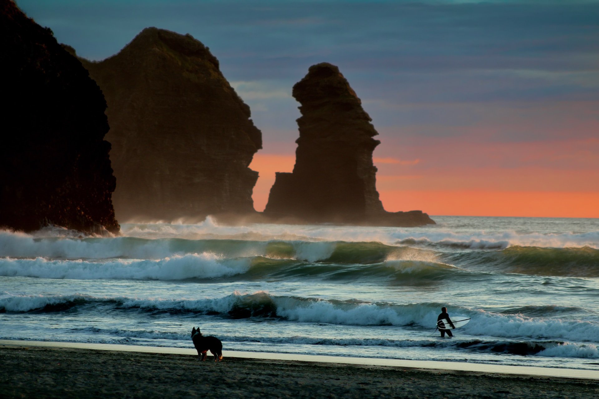 Piha Beach bij zonsondergang, met surfer en hond